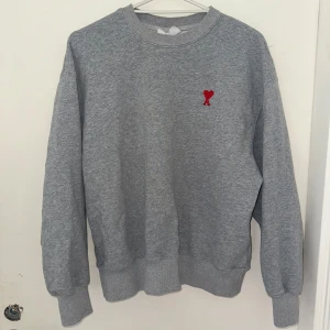 Grå sweatshirt från Ami Paris - Grå crewneck sweatshirt från Ami Paris med röd broderad hjärtlogga på bröstet. Klassisk passform med ribbade muddar vid ärmslut och midja. Perfekt basic med snygg detalj framtill, tillverkad i mjuk bomull.