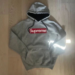 Grå Supreme hoodie med box logo - Supreme hoodie i grått med klassisk röd box logo på bröstet. Tröjan har huva med svarta snören, känguruficka och mjukt fleecefoder på insidan. Perfekt för dig som gillar streetwear och vill sticka ut med en ikonisk look.