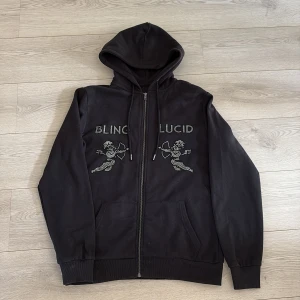 Svart hoodie med strass från Blinglucid - Svart hoodie från Blinglucid i storlek M med dragkedja och huva. Framsidan har strassdetaljer med texten 'BLING LUCID' och två änglar med pil och båge. Mjuk insida och ribbade muddar. Perfekt för dig som gillar bling och streetstyle. Använd fåtal gånger, hör av dig vid minsta fundering!