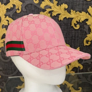 Rosa Gucci keps med GG-mönster - Snygg rosa keps från Gucci med klassiskt GG-monogram över hela kepsen. Grön och röd rand på sidan ger den ikoniska Gucci-looken. Justerbar rem bak i rosa läder. Perfekt för dig som vill sticka ut med en lyxig accessoar.