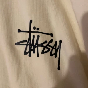 Vit Stüssy hoodie med logga - Snygg vit hoodie från Stüssy i storlek S. Tröjan har klassisk huva med snörning, stor svart Stüssy-logga på ryggen och mindre logga på bröstet. Tillverkad i mjuk bomull, perfekt för en chill streetwear-look.