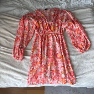 Blommig kort klänning från Gina Tricot - Superfin kort klänning från Gina Tricot i rosa, orange och vit med blommigt mönster. Klänningen har v-ringning, långa ärmar med resår och markerad midja. Perfekt för sommarens alla tillfällen.