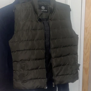 Grön dunväst från Stone Island - Säljer en mörkgrön dunväst från Stone Island med klassisk logotyp på ärmen. Västen har quiltad design, dragkedja framtill och två fickor. Perfekt för lager-på-lager och streetwear-stil. För liten för mig och har en liten reva på sidan där av priset 