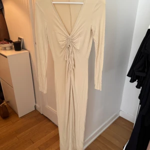 Beige långklänning med snörning - Säljer en beige långklänning med snygg v-ringning och snörning framtill som ger en cool rynkad effekt. Klänningen har långa ärmar och är figurnära i modellen. Perfekt för dig som gillar stilrena och trendiga plagg.