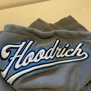 Grå hoodie från Hoodrich med brodyr - Säljer en grå hoodie från Hoodrich med fet broderad logga på huvan och coola tryck i vitt, blått och svart på bröstet och ärmarna. Hoodien har känguruficka och huva, perfekt för dig som gillar streetwear och vill sticka ut med snygga detaljer.