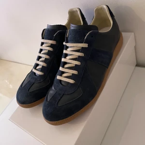 Maison Margiela Gats -  Säljer dessa feta Maison Margiela sneakers i mörkblå mocka och skinn med beige snörning och gummisula. Använda vid speciella tillfällen och finns inga tecken av defekter. Äktenhetsbevis finns. BARA box medföljer. Vid frågor/ funderingar är det bara att skriva.