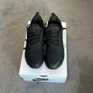 Nike Air Max 270 svarta sneakers - Nike Air Max 270 sneakers i helsvart färg med mesh-överdel och synlig Air Max-sula. Skorna har snörning och en modern, sportig look med rund tå och platt sula. Perfekt för dig som gillar streetstyle och vill ha bekväma sneakers med snygg design.