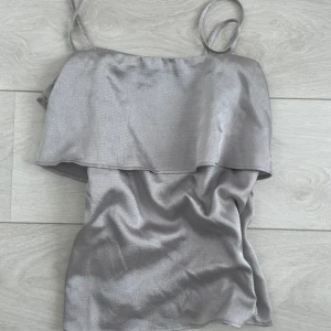 Silverfärgad linnetopp från H&M - Säljer en silverfärgad topp från H&M med tunna axelband och volangdetalj upptill. Toppen har en lätt skimrande yta och är perfekt för att lyfta en outfit med lite extra glans.
