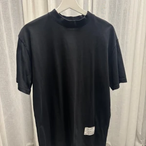 Svart t-shirt från Acne Studios - Svart t-shirt från Acne Studios med högre krage och korta ärmar. Tillverkad i 100% bomull och har en vit patch med logga nertill. 