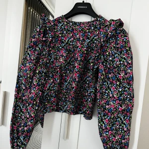 Blommig blus med volang från Zara, S - Supersöt blommig blus från Zara med volangdetaljer på axlarna och puffiga långärmar. Färgglada små blommor i rosa, blått, grönt och vitt mot svart botten. Perfekt för dig som gillar statement-plagg med mycket färg och mönster. Knappt använd i nyskick. Tröjan finns inte längre på Zara därför säljer jag den för 250kr då nypris var 399kr