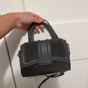 Svart handväska med pälsdetaljer - Snygg svart handväska med pälsliknande yta och detaljer i skinn. Väskan har ett stilrent handtag och en axelrem, perfekt för att lyfta din outfit. Designen är modern med en cool struktur och guldiga metalldetaljer på sidorna.