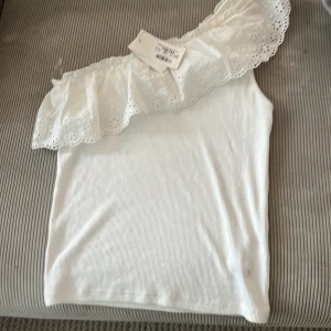 Vit offshoulder topp med volang från Kappahl  - Supersöt vit offshoulder topp från kappahl med bred broderad volang längs halsen. Toppen är ärmlös och har ett ribbat tyg i mjuk bomull. Volangen har små blommiga broderier.oanvänd tröja