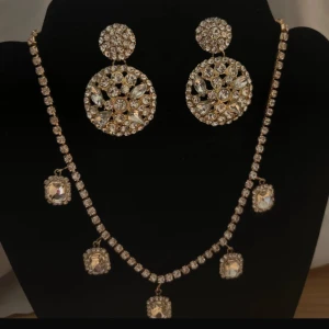 Glittrigt guldigt smyckeset med örhängen - Säljer ett lyxigt smyckeset med stora runda örhängen och matchande halsband i guldton. Båda delarna är dekorerade med glittrande stenar och gnistrande detaljer som verkligen drar blickarna till sig. Perfekt för dig som vill sticka ut med extra bling och glamour.