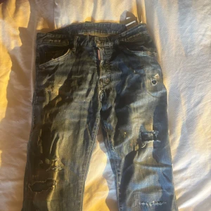 Blå slitna jeans från Dsquared2 - Säljer ett par blå jeans från Dsquared2 med coola slitningar och hål på benen. Jeansen har klassisk femficksdesign, låg midja och raka ben. Materialet är jeans i bomull och de har en schysst street vibe med detaljer som färgstänk och distressed look.