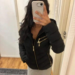 Svart pufferjacka från Hollies - INTRESSEKOLL! Svart pufferjacka från Hollies med guldiga dragkedjor och detaljer. Jackan har huva med snygg metallring och leopardmönstrat foder. Flera fickor med dragkedja framtill och quiltad design som ger en schysst form. 