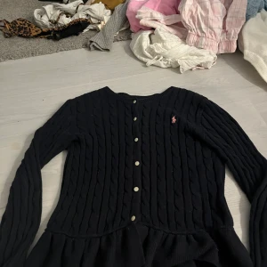 Svart kofta från Polo Ralph Lauren - Supersnygg svart kabelstickad kofta från Polo Ralph Lauren med klassiska knappar framtill och rosa broderad logga på bröstet. Långärmad modell med volangkant nedtill, perfekt för en preppy look. Gjord i mjuk bomull.