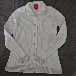 Vit ribbad kofta från Memories by Olé - Vit ribbstickad kofta med krage och knappar framtill från Memories by Olé. Långa ärmar och snygga beige ränder på muddar och nederkant. Klassisk och clean look, perfekt till jeans eller kjol. Skön stickad känsla och normal passform.