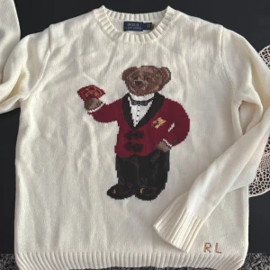 Vit stickad tröja Polo Bear Ralph Lauren - Vit stickad tröja från Polo Ralph Lauren med ikoniska Polo Bear-motivet framtill. Björnen har röd kavaj och håller spelkort. Rund halsringning, ribbade muddar och RL-broderi vid ärmslutet. Perfekt statement-piece för dig som gillar klassisk preppy stil.