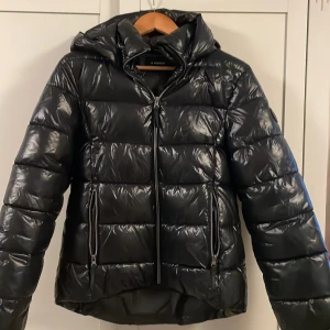 Svart pufferjacka från Everest - Säljer en svart, glansig pufferjacka från Everest med huva och dragkedja framtill. Jackan har två sidofickor med dragkedja och är quiltad för extra värme. Perfekt för kalla dagar och har en modern, loose passform som är enkel att styla med hoodies eller jeans.