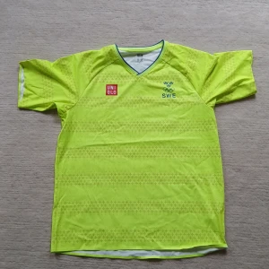 Svensk Uniqlo fotbollströja gul L - Gul fotbollströja från Uniqlo i storlek L, tillverkad i 100% polyester med Dry-Ex och Odor-Control teknik. Tröjan har korta ärmar, V-ringning med blå detalj och subtilt mönster över hela tyget. SWE och OS-logga på bröstet samt Uniqlo-märke.