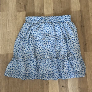 Blommig volangkjol från New yorker, M - Supersöt kjol med blått blommönster på vit botten från New yorker. Kjolen har volanger och resår i midjan för en bekväm passform. Perfekt för dig som gillar en romantisk och somrig stil.