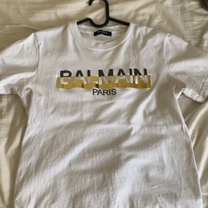 Vit Balmain t-shirt med logga - Vit t-shirt från Balmain med klassisk rund hals och tryckt logga i svart och gult på bröstet. Jätte bra skick! Pris kan diskuteras!