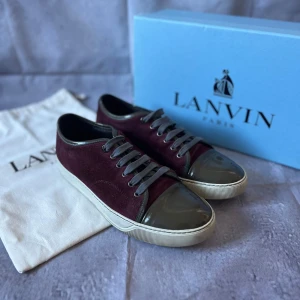 Lanvin Cap-Toe Paris Sneakers – Strl UK 7/ EU 41 – Vinröd mocka & lacktå – Med låda & dustbag - Säljer ett par Lanvin Paris Cap-Toe  i en unik kombination av vinröd mocka och olivgrön lacktå. Levereras med original dustbag och Lanvin-låda.   🎨 Färg: Vinröd mocka med olivgrön lacktå och vit yttersula 📏 Storlek: UK 7/ EU 41 📐 Mått: Innermått ca 26,5 cm (normal passform för EU 41) ♻️ Skick: 9/10, inga tecken på användning på framsidan eller lacktå. Lite smutsig sula. 🧵 Material: Mocka med lackad tå i skinn, gummisula, textilfoder och klassiska snören  ‼️Skickas dagen efter köpet genomförs‼️