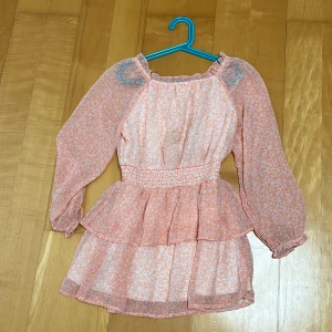 Ljusrosa blommig kortklänning - Supersöt ljusrosa kortklänning med små vita blommor, volangdetaljer och smock i midjan. Klänningen har långa, transparenta ärmar med resår vid handlederna och en rundad halsringning med lätt rynk. Perfekt för sommarens alla tillfällen!