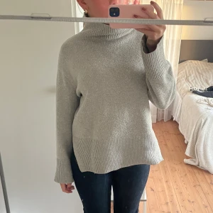 Ljusgrå polotröja stickad  - Mysig ljusgrå polotröja med breda ribbade muddar vid ärmslut och nederkant. Tröjan har en hög krage och är stickad i ett mjukt material. Passformen är lite loose och ärmarna är långa, perfekt för kyliga dagar.