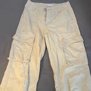 Beige cargobyxor från Bershka - Snygga beige cargobyxor från Bershka med flera stora fickor på sidorna och bak. Byxorna har normal passform, raka ben och är tillverkade i bomull. Perfekta för en avslappnad streetstil och passar till sneakers eller boots. Dock har de ingen lap för det står storleken men skulle tro att det är 34-36. 