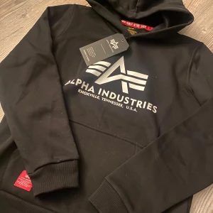 Svart hoodie från Alpha Industries - Svart hoodie från Alpha Industries med stor vit logga framtill. Klassisk huva, ribbade muddar och magficka. Materialet känns mjukt och skönt mot huden. Helt oanvänd och prislappen kvar.