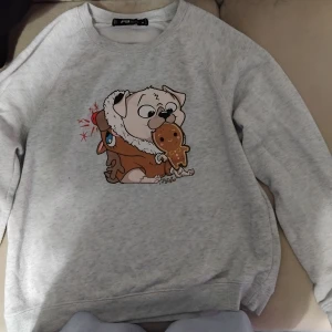 Grå sweatshirt med julmotiv FB Sister - Grå sweatshirt från FB Sister i storlek M med ett gulligt julmotiv framtill: en mops i tomteluva som äter en pepparkaka och en ren bredvid. Tröjan har rund hals och långa ärmar, perfekt för vinterns mysiga dagar.