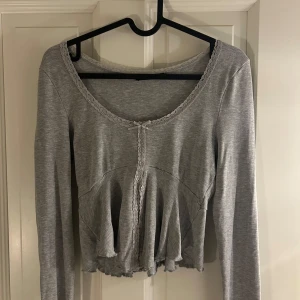 Grå croppad blus med spetsdetaljer - Supersöt grå croppad blus med långa ärmar och volang nertill. Spetskant runt halsringningen och framtill, plus en liten rosett som extra detalj. 