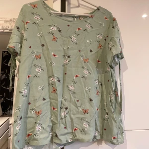 Grön blommig blus med fjärilar - Supersöt ljusgrön blus med kort ärm och knytband på sidorna. Blusen har ett blommigt mönster med små fjärilar och blommor i vitt, orange och rosa.                                                 Passar även dem som har M i storlek.