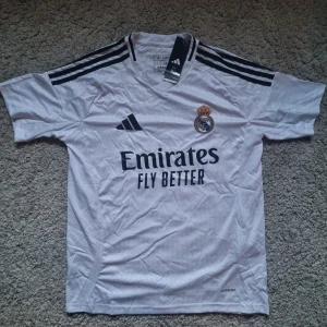 Real Madrid 25 Tröja - Snygg vit Real Madrid fotbollströja från Adidas med svarta detaljer och klubbmärke på bröstet. Tröjan har korta ärmar, V-ringning och klassiska Adidas-ränder på axlarna. Tillverkad i lätt och ventilerande AEROREADY-material. Perfekt för dig som älskar fotboll och Real.