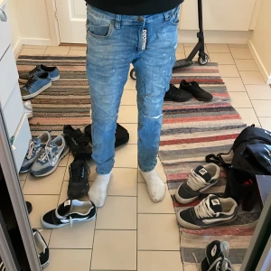 Blå jeans från Dsquared2, storlek 33 - Säljer ett par snygga ljusblå jeans från Dsquared2 med coola slitningar och stänkdetaljer. Jeansen har klassisk femficksdesign, knappgylf och tydliga sömmar. Passformen är slim och materialet är mjuk bomullsdenim. Perfekt för dig som gillar streetstyle och exklusiva märken.