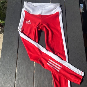 Röda Adidas leggings XS - Oanvända röda Adidas leggings med vita sidopaneler och klassiska tre ränder på benen. De har hög midja och dragkedja vid benslutet för extra stil och funktion. Perfekta för träning eller chil❣️