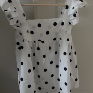 Vit prickig blus med volangärmar och rosett som knytning  - Supersöt vit blus med svarta prickar och volangärmar. Toppen har fyrkantig ringning, knytning i ryggen och lätt rynkade detaljer för en luftig känsla. Perfekt för dig som gillar lekfulla och trendiga plagg med extra detaljer. Aldrig någonsin använd.