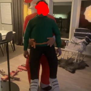 Uppblåsbar jultomte bär kostym - Rolig uppblåsbar kostym där det ser ut som att en jultomte bär dig på axlarna. Kostymen är i rött, grönt, svart och vitt och har inbyggd fläkt och batteridosa för att hålla den uppblåst. Perfekt för att sticka ut på julfesten eller maskerad!