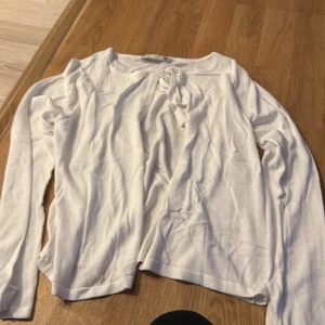 Vit långärmad tröja med knyt - En enkel och stilren vit långärmad tröja med knyt fram i halsen. Tröjan har en rak passform och är tillverkad i ett mjukt och tunt material, perfekt för lager-på-lager. Passar dig som gillar en clean och basic look.