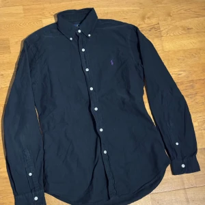 Svart skjorta från Ralph Lauren Slim - Svart skjorta från Ralph Lauren i slim fit med klassisk krage och vita knappar. Långärmad modell med lila broderad logga på bröstet. Skjortan är gjord i mjuk bomull och har en stilren, tidlös look som funkar till många olika outfits.