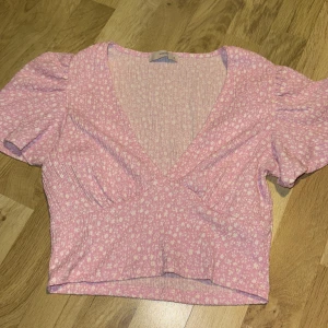 Rosa blommig topp från Pigalle - Säljer en söt rosa topp från Pigalle med blommigt mönster. Toppen har korta puffärmar och en v-ringad design som ger en feminin touch. Perfekt för vårens och sommarens varma dagar!