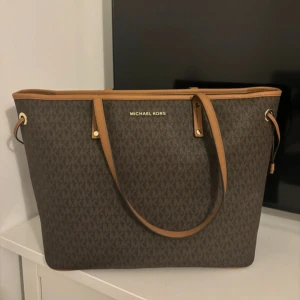 Brun axelväska från Michael Kors - Snygg brun axelväska från Michael Kors med klassiskt MK-mönster. Väskan har två handtag och detaljer i guld. Perfekt för att bära med sig allt man behöver under dagen. Stilren och praktisk design som passar till många tillfällen.