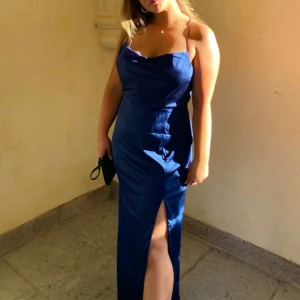 Marinblå långklänning med slits - Elegant marinblå långklänning med smala axelband och en stilren slits framtill. Perfekt för skolbal eller annan speciell tillställning. Klänningen har en figurnära passform som framhäver silhuetten.