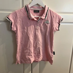 Rosa pikétröja från Kappa - Säljer en snygg rosa pikétröja från Kappa med korta ärmar och klassisk krage. Tröjan har svarta och vita detaljer på krage och ärmslut samt Kappa-logga på bröstet. Perfekt för en sportig look!
