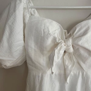 Vit klänning från Petal and Pup - Säljer denna vackra vit klänning från Petal and Pup/Whyte Valentyne. Puffiga off-shoulder ärmar och en söt knytdetalj framtill. Klänningen har en dragkedja bak och är i en  medelllång modell. Perfekt för sommarens alla tillfällen! Aldrig använd.