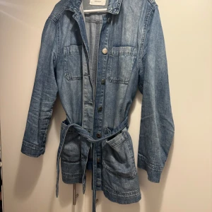 Blå jeansjacka med bälte - Snygg blå jeansjacka från Zara med knappar och bälte i midjan. Jackan har långa ärmar och två stora bröstfickor. 