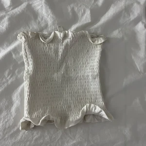 Vit smockad tubtopp - Säljer en söt vit smockad tubtopp med volangdetaljer upptill och nedtill. Perfekt för sommardagar och att matcha med jeans eller kjol. Toppen är elastisk och bekväm att bära.