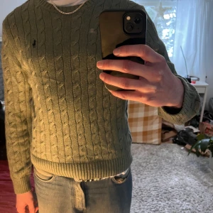 Grön stickad tröja från Ralph Lauren - Säljer en stilren grön stickad tröja från Ralph Lauren med kabelstickat mönster. Tröjan har långa ärmar och en broderad logotyp på bröstet. Perfekt för en klassisk och avslappnad look. En sämre sak är dock den nästintill osynliga fläcken i bild 5. Den syns inte när man bär den, om man inte kollar noga efter den. Jag är 195cm och väger 83kg, tycker den passar riktigt bra!