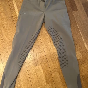 Beige ridbyxor från Beyond the Bit - Snygga beige ridbyxor från Beyond the Bit med greppande detaljer längs benen för extra stöd. Byxorna har en dragkedja framtill och bälteshällor. Perfekta för ridning med stil och komfort. Saknar knapp vid gylfen. 